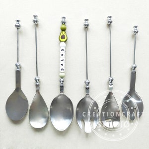 May include: Set of six silver salad serving utensils with long handles. The utensils include a spoon, a fork, and a serving spoon. One of the spoons has a beaded charm with the word "SALAD" spelled out in white letters.