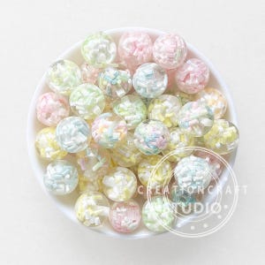 Puede incluir: Una colección de cuentas redondas y transparentes llenas de chispas de colores pastel. Las cuentas están en un cuenco blanco, con una variedad de colores que incluyen rosa, amarillo, verde y azul. Las cuentas son para manualidades.