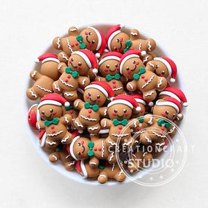 Pode incluir: Uma tigela branca cheia de pingentes de homem de gengibre. Os pingentes são marrons com detalhes de glacê branco, chapéus de Papai Noel vermelhos e gravatas borboleta verdes. O texto "CREATIONCRAFT STUDIO" é visível.