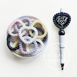 May include: A white bowl filled with iridescent heart-shaped bead charms. The charms are in various pastel colours, including pink, yellow, and purple. A black heart-shaped pen with the word "love" written on it is next to the bowl.