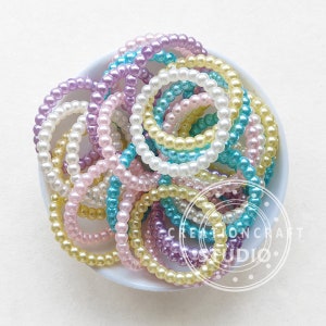 May include: A bowl filled with a variety of pastel-coloured pearl hair ties. The hair ties are made of small, round beads and come in shades of pink, yellow, blue, and purple.