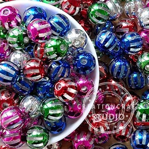 Puede incluir: Una colección de cuentas de colores y rayas en un cuenco blanco. Las cuentas son de varios colores, incluyendo rojo, azul, verde y rosa, con rayas plateadas. La imagen también contiene el texto "CREATIONCRAFT STUDIO".