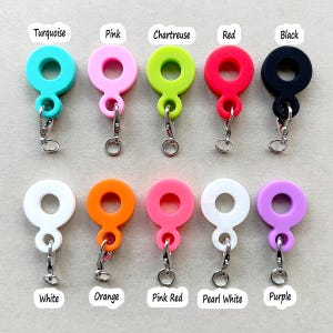 May include: Ten silicone charms in various colors, including turquoise, pink, chartreuse, red, black, white, orange, pink red, pearl white, and purple. Each charm features a double circle design with a silver metal loop.