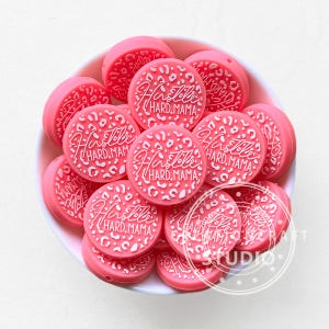 以下が含まれることがあります： ピンク色の丸いビーズのコレクション。ヒョウ柄のデザインと白い文字で「Hustle Hard Mama」と書かれています。ビーズは白いボウルに入っており、視覚的に魅力的な配置になっています。これらのビーズは、おそらく手作りのために使用されます。