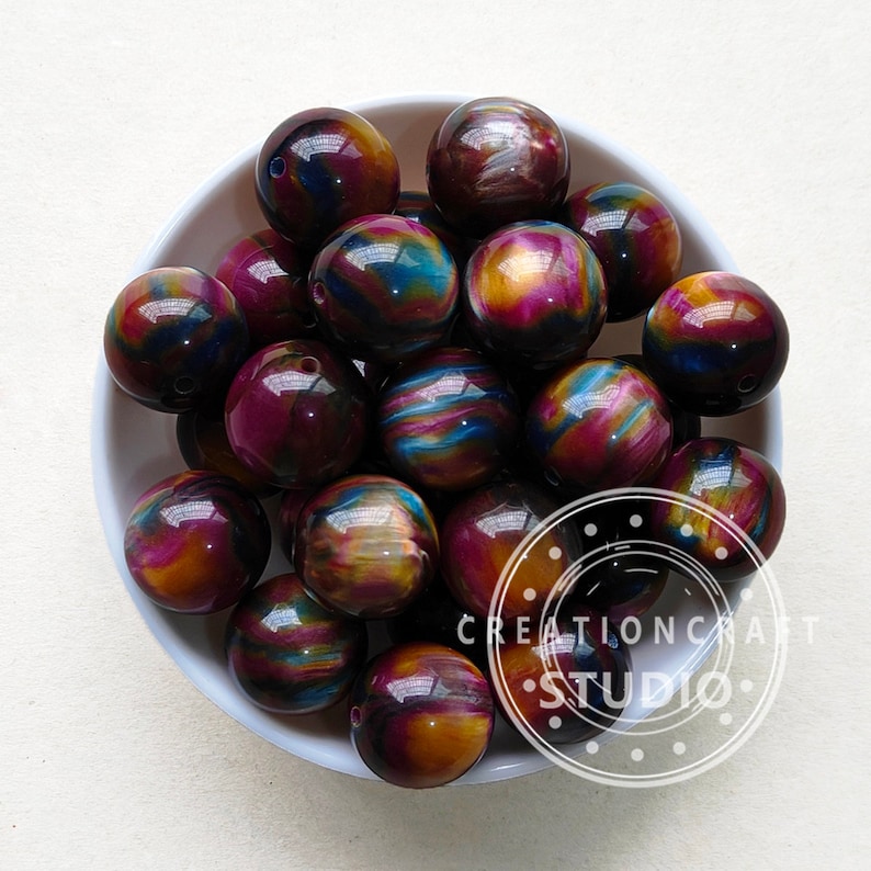 Pode incluir: Uma tigela de contas redondas iridescentes multicoloridas. As contas t&ecirc;m uma mistura de cores roxo, azul, dourado e preto.