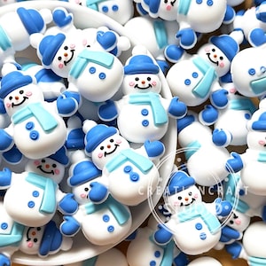 Könnte beinhalten: Eine Ansammlung kleiner, weißer Schneemann-Cabochons aus Kunstharz mit blauen Hüten, hellblauen Schals und zwei blauen Knöpfen am Körper. Jeder Schneemann hat eine orangefarbene Karottennase, schwarze Augen, einen lächelnden Mund und rosa Wangen.