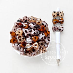 May include: A bowl of bow-shaped beads and a pen with bow-shaped beads. The beads are in shades of brown, tan, and white with a spotted pattern. The pen is white with silver accents. The beads are in a bowl.
