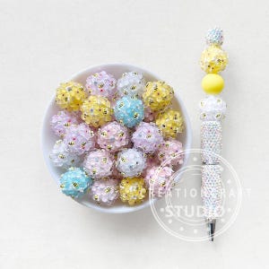 May include: A white bowl filled with colourful, round beads with tiny bee accents. A decorative pen with a rhinestone-covered barrel and yellow and white bead accents sits to the right. The pen has a silver tip.