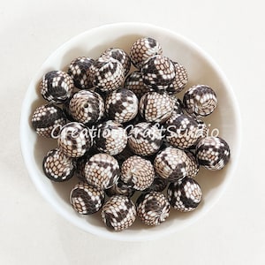 Puede incluir: Un bol de cuentas de silicona marrones y blancas con un patrón de piel de serpiente. Las cuentas son redondas y tienen una superficie lisa.