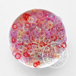 Könnte beinhalten: Eine weiße Schüssel gefüllt mit farbenfrohen, durchscheinenden Perlen. Die Perlen sind ringförmig mit einer strukturierten Oberfläche und in den Farbtönen Rot, Rosa, Lila und Gelb gehalten. Die Perlen sind in der Schüssel verstreut und erzeugen eine lebendige Darstellung.