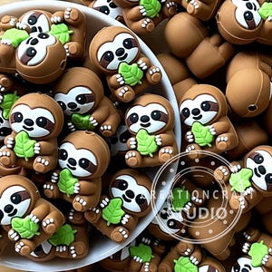 Pode incluir: Uma coleção de pingentes de preguiça marrons, cada um segurando uma folha verde. As preguiças têm rostos brancos, olhos pretos e contornos marrons. Os pingentes estão em uma tigela branca e espalhados. Ideal para artesanato ou joalheria.