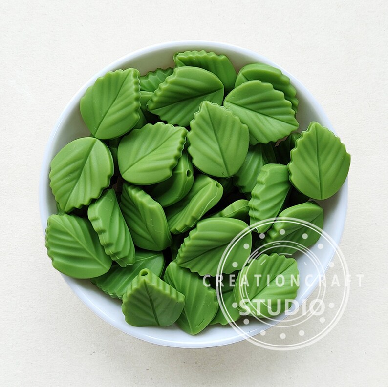 May include: A white bowl overflowing with green leaf-shaped beads. Each bead features detailed veining and a glossy finish. The leaves are a vivid green, evoking a natural aesthetic. The image is well-lit, accentuating the texture and form of the beads. The beads are likely for crafting.