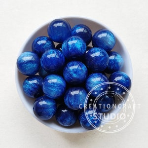 May include: A bowl filled with blue and silver glitter resin beads. The beads are round and have a shimmery finish.