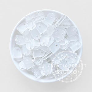 May include: A collection of small, translucent plastic clips with a rounded top and a hole, displayed in a white bowl. The clips are evenly distributed, suggesting a craft or DIY supply.