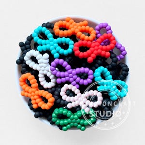 Pode incluir: Uma tigela branca cheia de contas coloridas em forma de laço. As contas são de várias cores, incluindo laranja, vermelho, roxo, azul, branco, rosa e verde. A tigela é cercada por contas pretas.