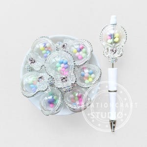 May include: A collection of clear, skull-shaped containers filled with colorful beads, arranged on a white plate. A white pen with a skull-shaped top, also filled with beads, is positioned to the right. The pen has the text "CREATION CRAFT STUDIO".
