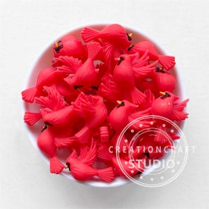 May include: A bowl filled with bright red cardinal bird charms. The charms are made of a smooth, glossy material and have black eyes and beaks.