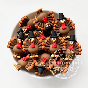 Puede incluir: Una colección de adornos con forma de pavo marrón con plumas de cola naranjas y marrones, carúnculas rojas y sombreros de copa negros. Los pavos están en un cuenco blanco. El texto "CREATIONCRAFT STUDIO" es visible.