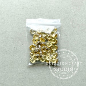 May include: A clear plastic bag filled with gold-colored, flower-shaped beads and silver-colored rhinestone spacers. The beads have a textured surface and are likely used for jewelry making or other crafts. The bag is on a light gray background.