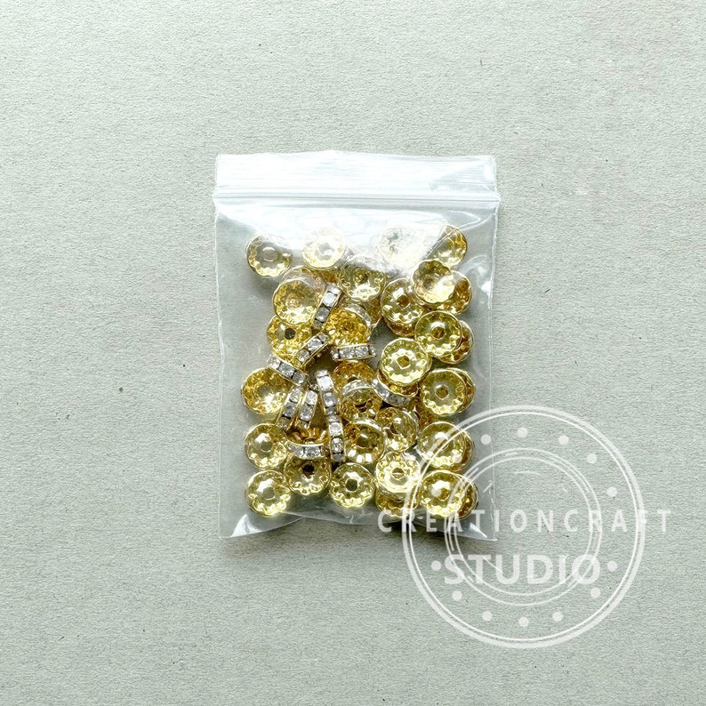 May include: A clear plastic bag filled with gold-colored, flower-shaped beads and silver-colored rhinestone spacers. The beads have a textured surface and are likely used for jewelry making or other crafts. The bag is on a light gray background.