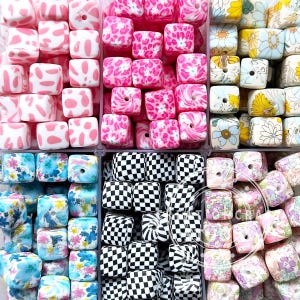 Peut inclure: Une collection de perles carrées aux motifs et couleurs variés. Les perles comprennent des pois roses et blancs, des imprimés animaliers roses, des damiers noirs et blancs, des motifs floraux et des motifs tourbillonnants. Ces perles sont probablement destinées à la fabrication de bijoux.