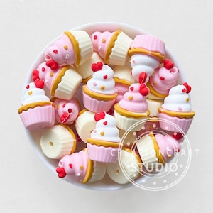 Peut inclure: Un bol blanc rempli de breloques miniatures en forme de cupcake. Les cupcakes sont roses et blancs avec des bases jaunes et des accents de cœur rouges. Le texte "CREATIONCRAFT STUDIO" est visible.