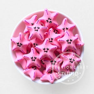 May include: A bowl filled with pink starfish beads, each with a smiling face and purple detailing. These decorative beads are likely for crafting or jewellery making. The image includes the text "CREATIONCRAFT STUDIO".