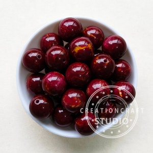 Op de afbeelding: Een kom gevuld met bordeauxrode en goudmarmerende ronde kralen. De kralen zijn glad en glanzend.