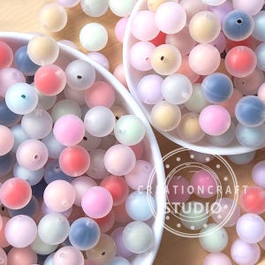 Puede incluir: Primer plano de dos cuencos blancos llenos de cuentas redondas y coloridas en tonos pastel de rosa, azul, amarillo y blanco. Las cuentas están dispersas, con algunos agujeros visibles. La imagen incluye el texto "CREATIONCRAFT STUDIO".