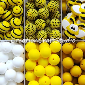 Pode incluir: Um conjunto de contas de silicone em três cores: branco, amarelo e amarelo mais escuro. As contas estão dispostas em três filas, com as contas brancas na primeira fila, as contas amarelas na segunda fila e as contas amarelas mais escuras na terceira fila. Também há algumas contas de silicone com tema de abelha na imagem.