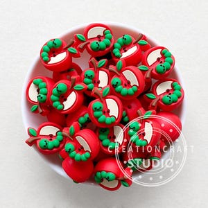May include: A bowl filled with red and green apple-shaped silicone charms with a worm inside each apple. The charms are for jewellery making or other craft projects.