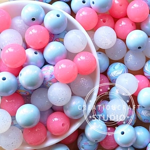Puede incluir: Una colección de cuentas redondas de colores en tonos rosa, azul y blanco. Algunas cuentas tienen un acabado brillante, otras son sólidas. Las cuentas están esparcidas en un cuenco blanco y alrededor. Las cuentas son para manualidades.