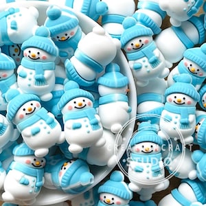 May include: A collection of small, white snowman figurines with blue hats, scarves, and accents. Each snowman has a smiling face with rosy cheeks and a carrot nose. The snowmen are scattered in a white bowl, creating a festive winter scene.