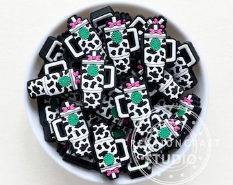 May include: A collection of black and white cow-print coffee cup charms with pink bows and green accents, piled in a white bowl. The charms have a handle and a logo. The charms are likely for crafting or jewelry making.