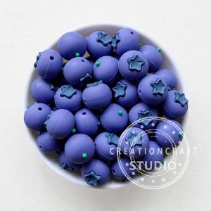 May include: A bowl filled with purple silicone beads shaped like blueberries. Each bead has a green star on top.