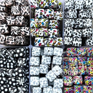 May include: A selection of square beads with diverse patterns and colours. Designs include leopard print, polka dots, and abstract swirls in black, white, grey, and vibrant hues. The beads are arranged in clear plastic containers.