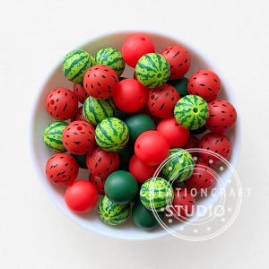 May include: A white bowl filled with various beads. The beads are red, green, and watermelon-themed, with some solid colors and others featuring a watermelon rind design. The image includes the text "CREATION CRAFT STUDIO".