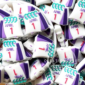 May include: A collection of white, purple, and teal calendar charms, each displaying "APRIL 1". The charms have a 3D design with a calendar shape, featuring a teal spiral binding and a red "1" on the date.