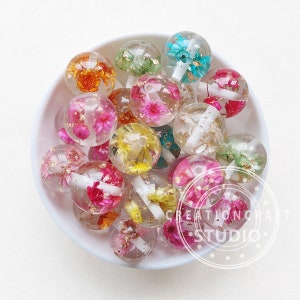 Pode incluir: Uma tigela cheia de contas de resina transparente, cada uma contendo flores prensadas e folha de ouro. As contas são de várias cores, incluindo rosa, amarelo, laranja, verde e azul.