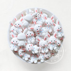 Puede incluir: Un cuenco lleno de figuras de conejitos blancos. Cada conejito tiene orejas forradas de rosa, mejillas rosadas y un lazo azul. Algunos conejitos están de pie, otros tumbados. Las figuras son pequeñas y parecen estar hechas de un material liso.