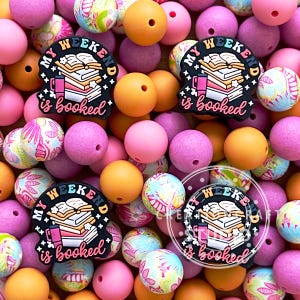 May include: An assortment of colorful beads in shades of pink, orange, and patterned designs. Several black charms with the text "My Weekend is booked" and a book and mug graphic are scattered throughout the image.