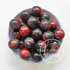 Pode incluir: Uma tigela de contas redondas de resina coloridas com redemoinhos de vermelho, verde e dourado. As contas t&ecirc;m um di&acirc;metro aproximado de 2,5 cm.