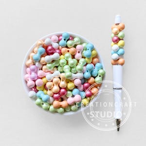May include: A white pen adorned with colourful, rounded beads in shades of orange, yellow, pink, and blue. A white bowl filled with similar beads sits next to the pen. The beads are in various pastel colours.
