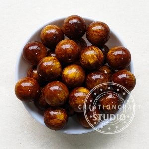 Pode incluir: Uma tigela cheia de 18 contas redondas, marrons e douradas com um padr&atilde;o de espiral. As contas s&atilde;o brilhantes e t&ecirc;m um brilho met&aacute;lico.