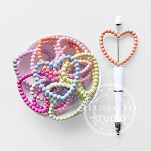 May include: A collection of colorful beaded bracelets in a white bowl, alongside a white pen with a heart-shaped beaded top. The bracelets are in shades of pink, purple, blue, yellow, and orange. The pen has a silver tip and a peach-colored heart.