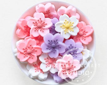 Puede incluir: Un bol lleno de encantos de flores de silicona de colores en rosa, morado y blanco. Los encantos tienen forma de flores de cerezo y tienen un efecto 3D.