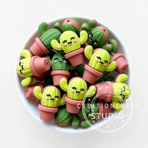 Pode incluir: Uma coleção de contas em forma de cacto sorridentes numa tigela branca. As contas são principalmente verdes e amarelas, com vasos cor-de-rosa. O texto "CREATIONCRAFT STUDIO" é visível. Ideal para artesanato.