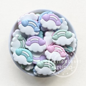 Puede incluir: Un bol lleno de abalorios de silicona de color pastel con forma de nubes con arcoíris y corazones. Los abalorios son perfectos para añadir un toque de fantasía a joyas, llaveros u otras manualidades.