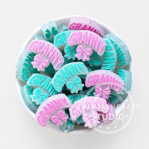 Pode incluir: Uma coleção de enfeites de resina com tema Granny em uma tigela branca. Os enfeites têm a forma de um semicírculo com a palavra "GRANNY" em rosa ou turquesa e um laço. O fundo é branco.