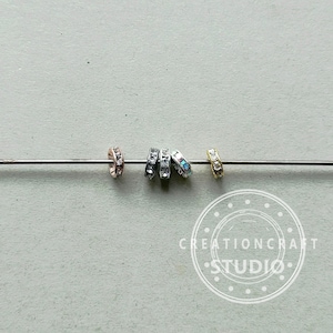 May include: Close-up of several small, decorative ring beads in various metallic finishes: rose gold, silver, and gold. Each ring is encrusted with small, clear crystals. The beads are strung on a thin, silver wire against a neutral gray background.